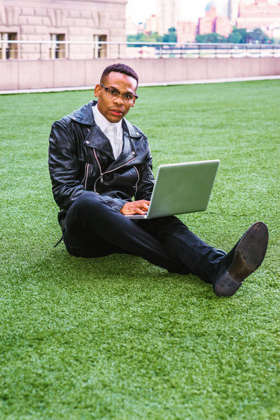 African American college student studying in New York. Wearing leather jacket, jeans, leather shoes, glasses, a young black man sitting on green lawn on campus, reading, working on laptop computer. - Fotoğraf, Görsel