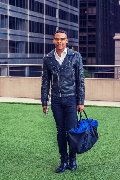 Man Urban Casual Fashion. Wearing black leather jacket, jeans, leather shoes, glasses, holding laptop computer, carrying duffel bag, African American college student walking on campus in New York. - Фото, изображение