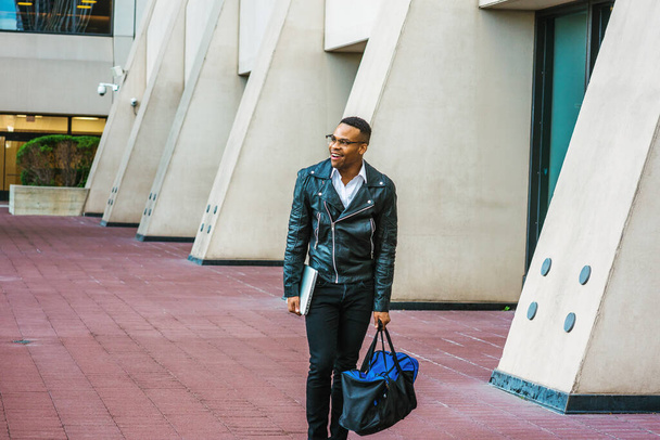 Man Urban Casual Fashion. Wearing black leather jacket, jeans, glasses, holding laptop computer, carrying duffel bag, happy African American college student walking on street in New York, smiling. - Foto, Imagen