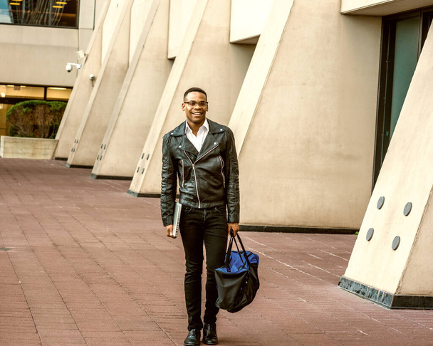 Homem Urbano Moda Casual. Vestindo jaqueta de couro preto, jeans, sapatos de couro, óculos, segurando computador portátil, levando mochila, estudante universitário afro-americano andando na rua em Nova York. - Foto, Imagem