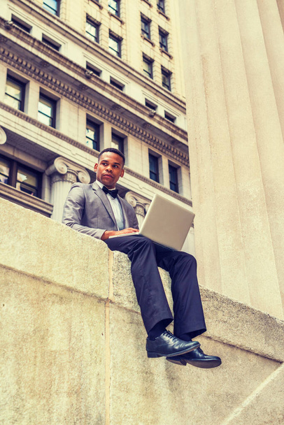 Hombre de negocios afroamericano que trabaja en Nueva York, vistiendo chaqueta gris, pajarita, pantalones negros, zapatos de cuero, sentado en la calle, leyendo, trabajando en la computadora portátil, pensando. Efecto filtrado. - Foto, imagen