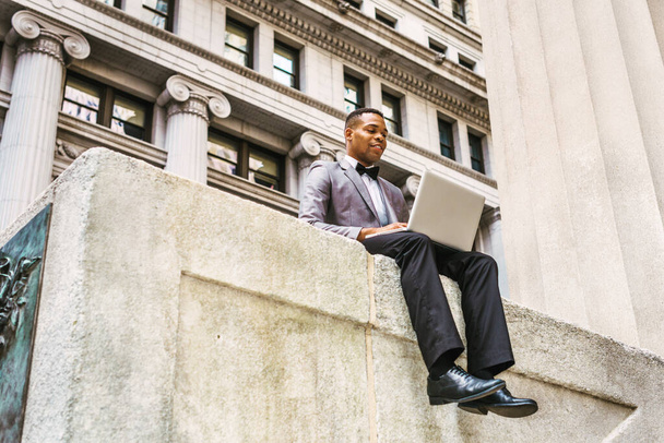 African American businessman working in New York, wearing gray blazer, bow tie, black pants, leather shoes, siting on street, reading, working on laptop computer, thinking. Filtered effect. - Photo, image