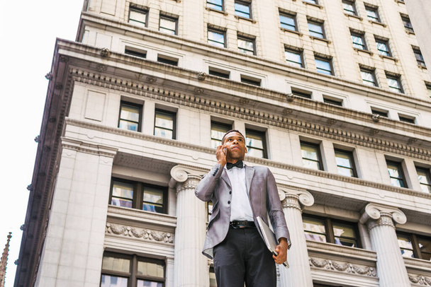 Puissance de la technologie. Homme d'affaires afro-américain travaillant à New York, vêtu d'un blazer gris, nœud papillon, debout dans la rue, tenant un ordinateur portable, appelant sur un téléphone portable. Effet filtré. - Photo, image