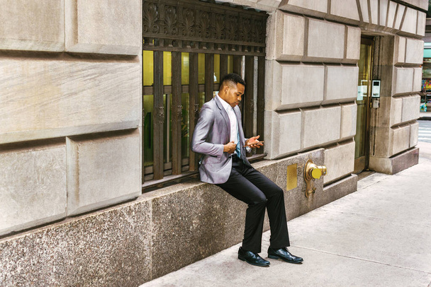 Homem afro-americano lendo mensagem no celular na rua, viajando, trabalhando em Nova York, vestindo blazer cinza, calças pretas, sapatos de couro, sentado na moldura da janela do vintage. Olhar filtrado. - Foto, Imagem