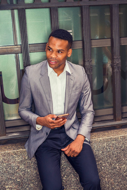 Sms 'en overal. Afro-Amerikaanse man draagt grijze blazer, wit ondershirt, zwarte broek, zittend op vintage stijl raamkozijn op straat in New York, mobiele telefoon vasthoudend. Gefilterd effect. - Foto, afbeelding