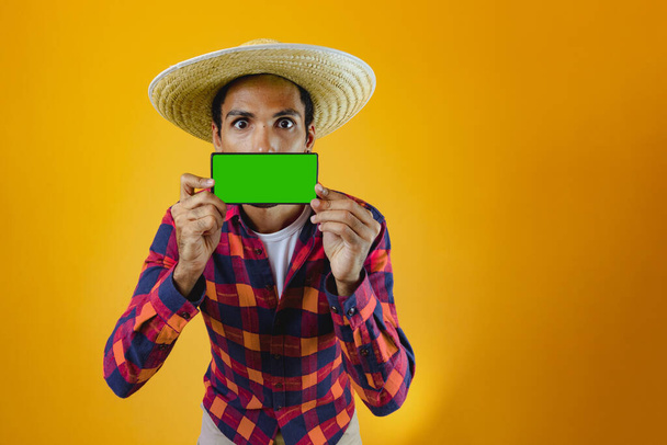 Black Man With Junina Party Outfit holding a Mobile in Front of a mouth Isolated on Yellow Background. Young man wearing traditional clothes for Festa Junina - Brazilian June festival. - Photo, Image