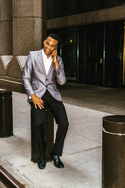 Αφροαμερικανός επιχειρηματίας που εργάζεται στη Νέα Υόρκη. Φορώντας γκρι σακάκι, μαύρο παντελόνι, δερμάτινα παπούτσια, ένας μαύρος να κάθεται στο δρόμο έξω από το γραφείο, χαμογελώντας, μιλώντας στο κινητό τηλέφωνο. Φιλτραρισμένο αποτέλεσμα. - Φωτογραφία, εικόνα
