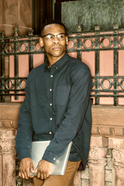 Αφροαμερικάνος φοιτητής που σπουδάζει στη Νέα Υόρκη, φορώντας μπλε πουκάμισο, καφέ παντελόνι, γυαλιά, κρατώντας φορητό υπολογιστή, στέκεται ενάντια σε vintage στυλ τοίχο στο δρόμο, σκέψης. Χρώμα φιλτραρισμένο εφέ. - Φωτογραφία, εικόνα