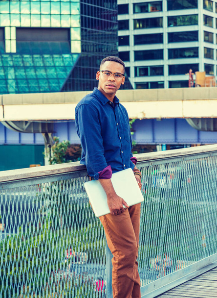 Un jeune Afro-Américain a grandi à Big City. Portant chemise bleue, lunettes, tenant un ordinateur portable, un étudiant noir debout contre la clôture dans le quartier des affaires avec des bâtiments élevés, pensant. - Photo, image