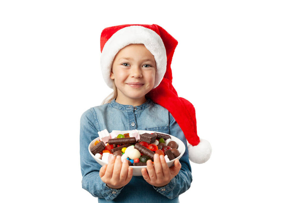 Cute little girl in santa hat with a plate of sweets isolated on white background. Happy child with lollipops and chocolate candies in hands. - Foto, imagen