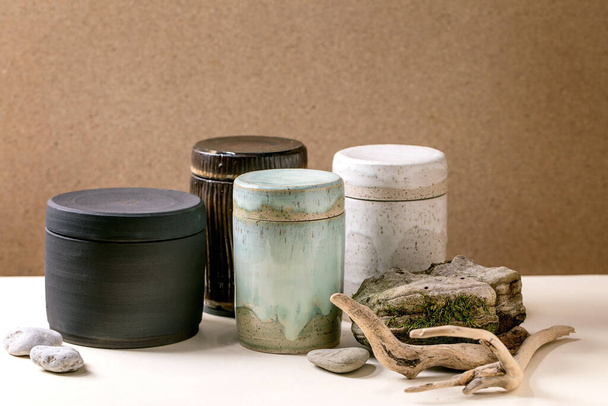 Set of closed craft ceramic jars with lids, different size and colors, with dry branch and stones standing on table. Brown wall, copy space - Fotó, kép