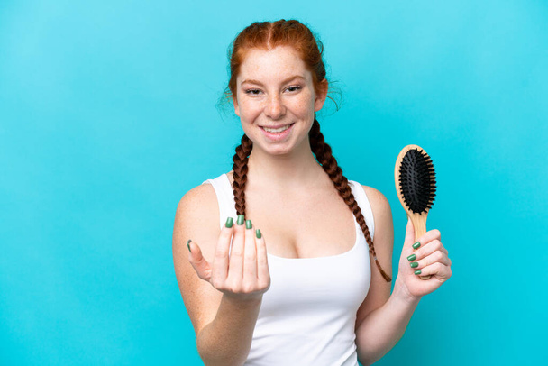 Young caucasian reddish woman with hair comb isolated on blue background inviting to come with hand. Happy that you came - Foto, immagini
