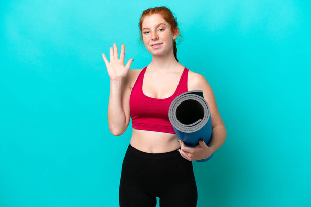 Junge Sportlerin geht zu Yoga-Kursen und hält eine Matte isoliert auf blauem Hintergrund und grüßt mit fröhlichem Gesichtsausdruck - Foto, Bild