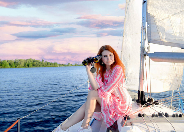 Beautiful red-haired girl in a pink mini dress sitting on a yacht with binoculars. The background is a stunning pink sunset, sky, white sails, sea. Summer. Travel concept. Copy space on the left - Foto, imagen