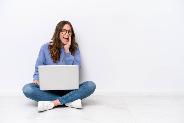 jonge Kaukasische vrouw met een laptop zitten op de vloer geïsoleerd op witte achtergrond schreeuwen met mond wijd open - Foto, afbeelding