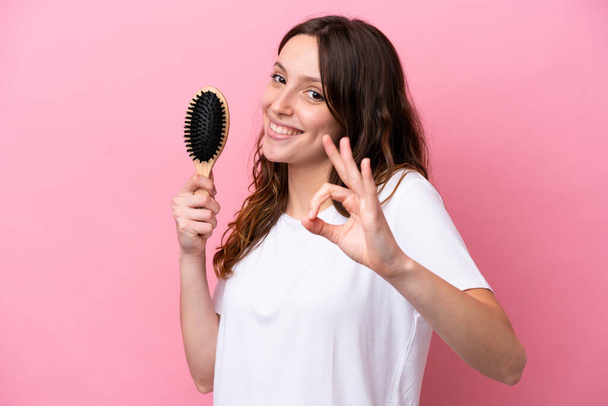 Young caucasian woman with hair comb isolated on pink background showing ok sign with fingers - Valokuva, kuva