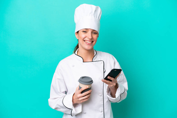 Молодая кавказская шеф-повар женщина изолирована на синем фоне держа кофе, чтобы забрать и мобильный - Фото, изображение