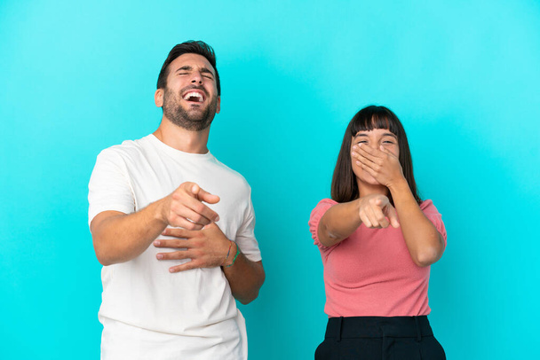 Молодая пара изолирована на синем фоне, указывая пальцем на кого-то и много смеясь - Фото, изображение