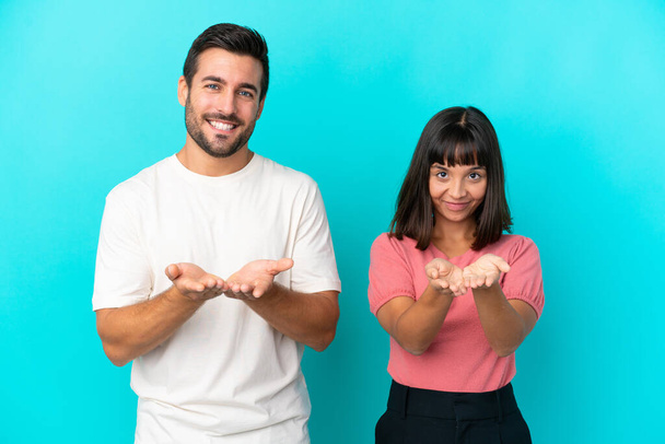Young couple isolated on blue background holding copyspace imaginary on the palm to insert an ad - Foto, Imagen