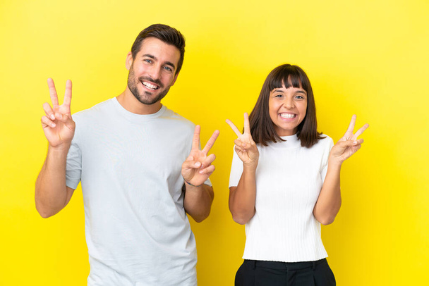 Young couple isolated on yellow background smiling and showing victory sign with both hands - Fotografie, Obrázek