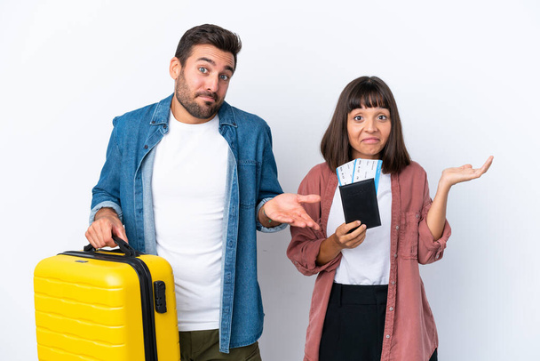 Young traveler couple holding a suitcase and passport isolated on white background having doubts while raising hands and shoulders - Valokuva, kuva