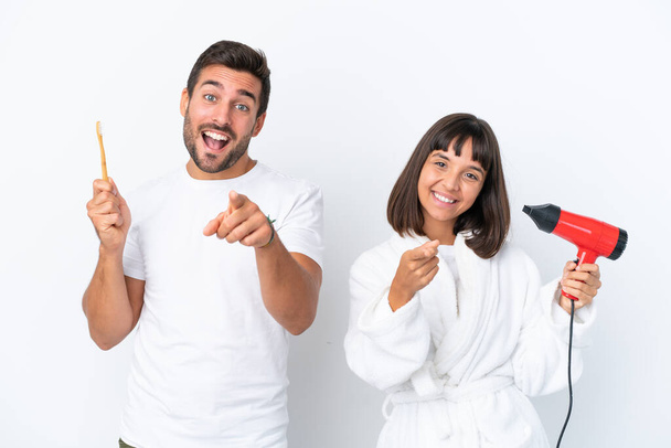 Jovem casal caucasiano segurando um secador de cabelo e escova de dentes isolado no fundo branco aponta dedo para você com uma expressão confiante - Foto, Imagem