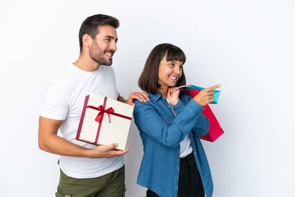 Jeune couple tenant des sacs à provisions et présent isolé sur fond blanc pointant vers le côté pour présenter un produit - Photo, image