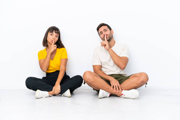 Young couple sitting on the floor isolated on white background showing a sign of closing mouth and silence gesture - 写真・画像