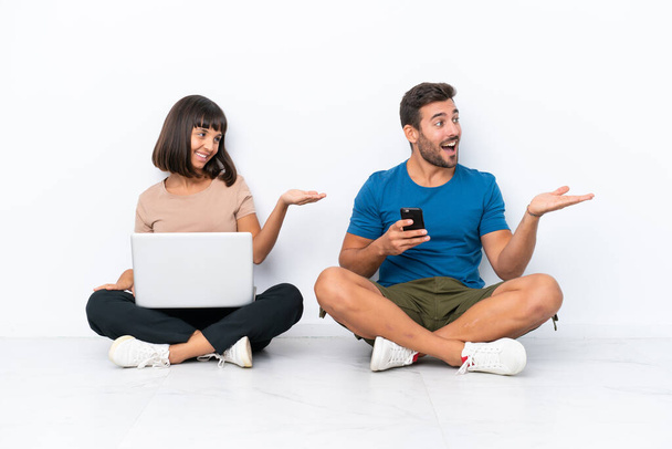 Giovane coppia seduta sul pavimento in possesso di pc e telefono cellulare isolato su sfondo bianco che punta indietro e presenta un prodotto - Foto, immagini