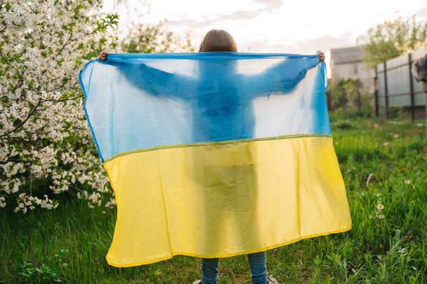Ukrainian flag and child silhouette. Silhouette of a child behind a Ukrainian flag, illuminated by the afternoon sun in a blue and yellow portrait. - Φωτογραφία, εικόνα