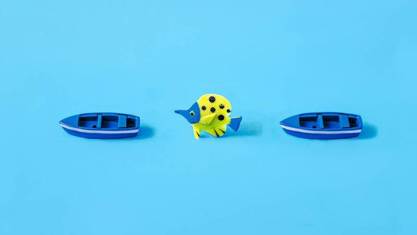 A blue and yellow fish between two blue boats against pastel blue background. Minimal creative concept for summer holidays or travel agency advertisement. Surreal design. Copz space - Photo, Image