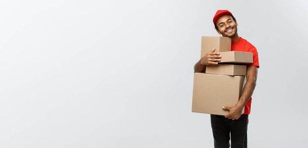 Αντίληψη παράδοσης - Πορτρέτο του Happy African American delivery man σε κόκκινο πανί κρατώντας ένα πακέτο κουτί. Απομονωμένος στο Grey studio Background. Αντιγραφή χώρου. - Φωτογραφία, εικόνα