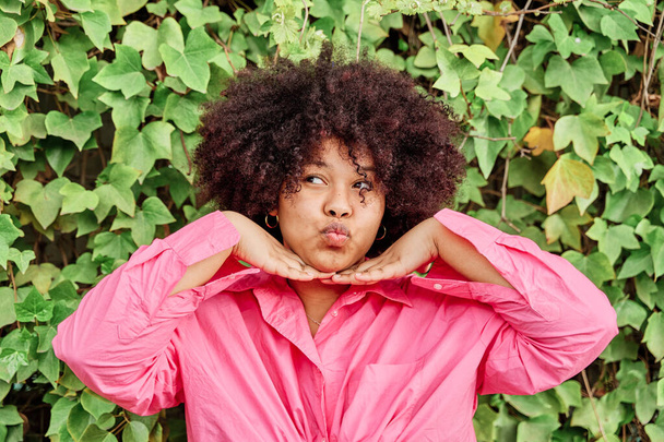 african american woman with curly hair posing to camera on background of plants. portrait of woman with comical gesture, sending kiss holding cheeks in hands - Photo, Image