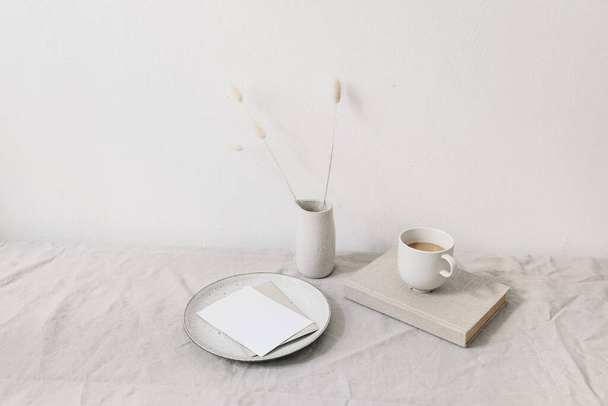 Neutral breakfast still life scene. Blank greeting card, ceramic plate. Vase with dry lagurus bunny tail grass on book. Cup of coffee on book. Linen table cloth, white wall. Scandinavian interior. - Fotó, kép