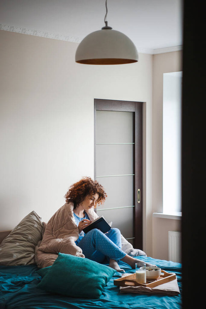 Portrét kavkazské kudrnaté ženy, která sedí na posteli, jí zdravou snídani a čte zajímavou knihu. Čtení ranních knih. Odpočinek, pohodlí, volný čas - Fotografie, Obrázek