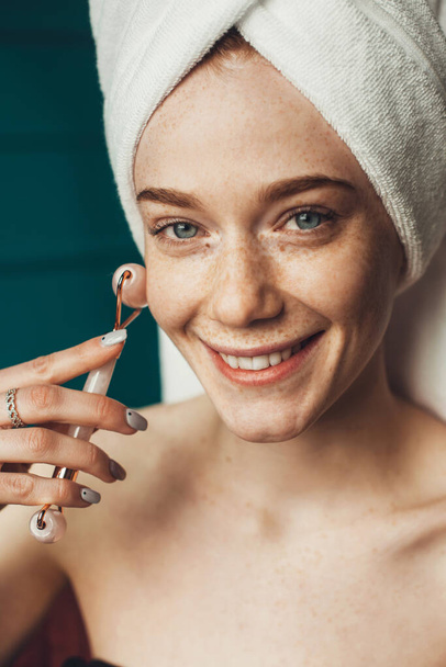 Woman with freckles wearing towel on head using a jade face roller with natural quartz stones. Smiling happy face. Beautiful young girl. Beauty face. Health - Photo, image