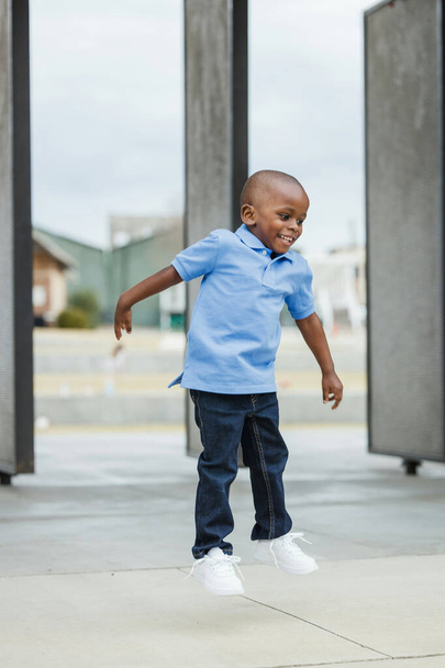 A cute little boy of preschool age with a blue shirt downtown in the city in denim jeans jumping up and down and playing - Foto, immagini