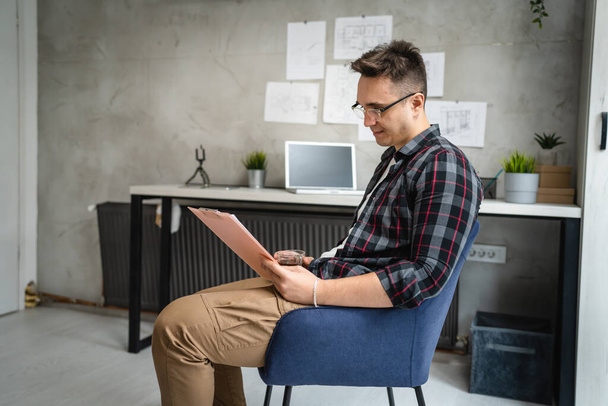 ένας άνδρας νεαρός καυκάσιος άνδρας κάθεται δίπλα στο γραφείο στη δουλειά στα έγγραφα μελέτης γραφείου χαλαρώνοντας ενώ κρατά διάγραμμα κλιπ με χαρτιά πραγματικοί άνθρωποι αντιγράφουν χώρο αρσενικό μηχανικό σχεδιασμό έργου - Φωτογραφία, εικόνα