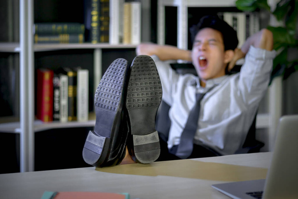 Shoes on table, Asian young tired staff officer man feeling exhausted and sleepy after working hard overtime at night, resting his feet on desk and take a nap after finishing work. - 写真・画像