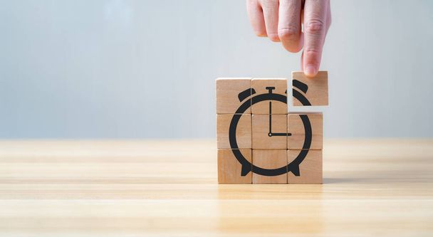 dłoń biznesmena trzymając i montując drewnianą kostkę z ikoną budzika w kształcie zegara. Koncepcja oszczędzania czasu na pracę w celu zmniejszenia kosztów. - Zdjęcie, obraz