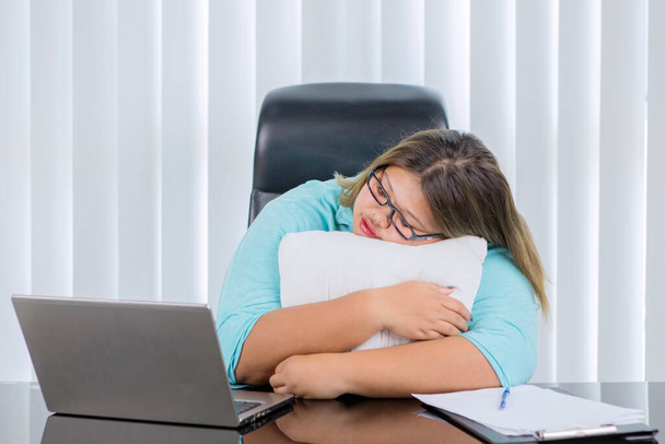 Mulher de negócios gorda cansada abraçando um travesseiro enquanto dormia na mesa no escritório com um laptop na mesa - Foto, Imagem