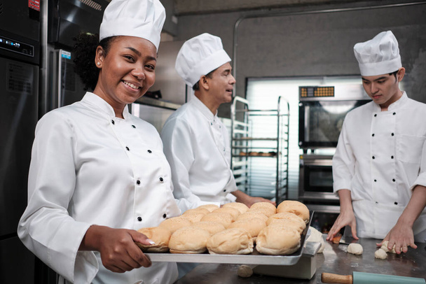 Portrait of African American female chef in white cooking uniform looking at camera with cheerful smile and proud with tray of bread in kitchen, pastry foods professional and fresh bakery occupation. - 写真・画像