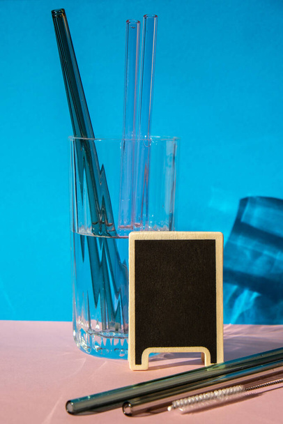 Επαναχρησιμοποιήσιμα γυάλινα καλαμάκια σε γυαλί με νερό σε πολύχρωμο φόντο με ετικέτα για το κείμενό σας Τρόπος ζωής Χαμηλή σπατάλη Οικολογικό Άνθος Ποτού Σετ με βούρτσα καθαρισμού. Μηδέν απόβλητα, πλαστική ελεύθερη έννοια - Φωτογραφία, εικόνα