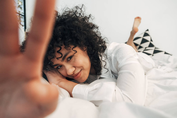 Mujer tímida de raza mixta joven mirando a la cámara acostada en la cama que cubre la lente con la mano. Enfoque selectivo en la cara. - Foto, imagen