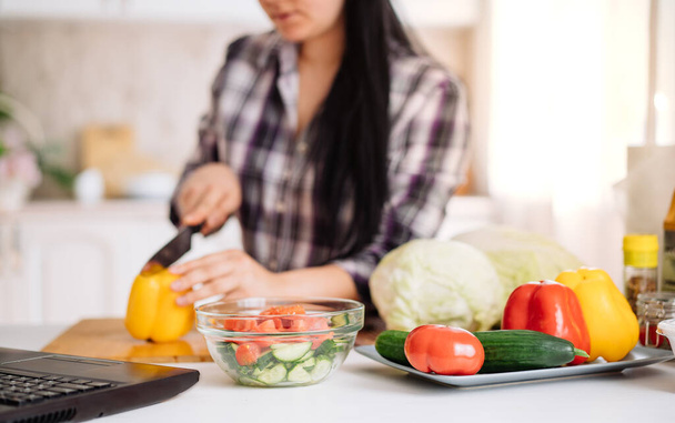 Eine fröhliche junge Frau kocht in der Küche mit einem Online-Publikum. Das Mädchen bereitet einen Salat zu, schneidet während einer Online-Sendung eine Gurke in Scheiben. Online kochen. Selektiver Fokus - Foto, Bild