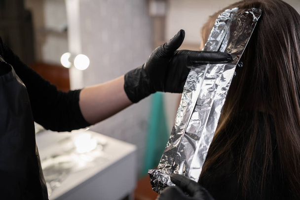 Close-up of hands of hairdresser hiding strands of hair of beautiful young woman with aluminum foil by changing hair color in spa salon - 写真・画像