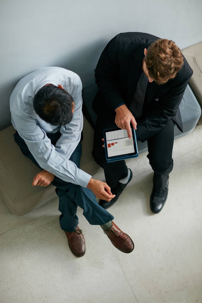 Переходимо до щоденних чисел. Високий кут зйомки двох бізнесменів, які розмовляють разом через цифровий планшет, сидячи в сучасному офісі
. - Фото, зображення