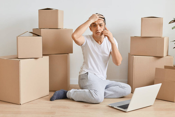 Image of Caucasian shocked man sitting on floor with laptop and talking on mobile phone among cardboard boxes, having problems with his work, looking at camera, forgot something important. - Фото, изображение