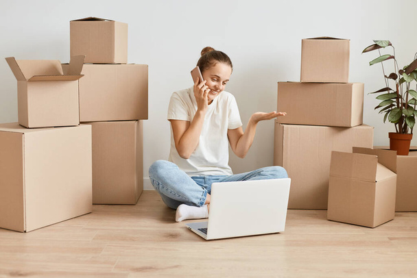 Image of confused dark haired woman wearing jeans and white t shirt sitting on floor with crossed legs among carton boxes and talking phone, being not sure, working on laptop. - Fotó, kép