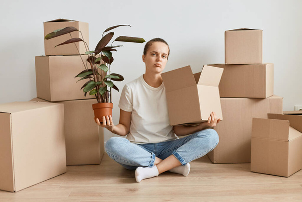 Sad upset woman in white t shirt and jeans sitting on floor in lotus pose, looking at camera, being surrounded with cardboard boxes, holding flower pot with plant, looking at camera with pout lips. - Fotografie, Obrázek
